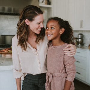 A white woman with long brown hair wearing a blouse and green slacks hugging her daughter with mocha skin toned excited about the report card
